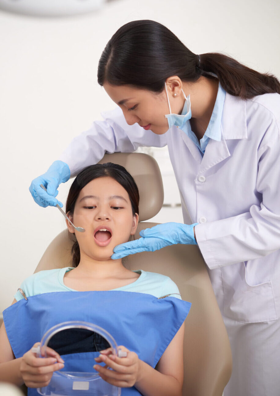 asian patient sitting with open mouth holding mirror dentist checking her teeth scaled e1752596670403